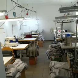 Wnętrze szwalni z maszynami do szycia, stosami materiałów w odcieniach szarości i beżu, oświetleniem jarzeniowym oraz widocznymi kablami pod sufitem.