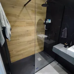 Nowoczesna kabina prysznicowa z drewnopodobnymi i heksagonalnymi płytkami, czarną armaturą i szklaną ścianką. Uwagę zwraca nietypowe połączenie kolorów i materiałów.