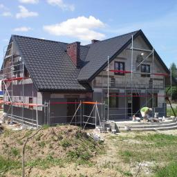 REMOS Piotr Dobkowski - Dom jednorodzinny w trakcie ocieplania, widoczne rusztowania, warstwa szarego ocieplenia, pracownicy oraz przygotowane materiały budowlane i narzędzia.