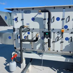 Centrala wentylacyjna z rurami w izolacji, manometrem, elementami sterującymi i przewodami na dachu budynku, widok z boku.