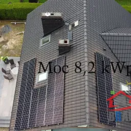 Widok z góry na dom z ciemną dachówką i panelami słonecznymi o mocy 8,2 kWp, w tle zaparkowane samochody i altanka.