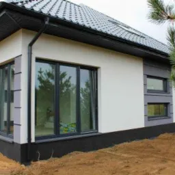 Nowoczesny dom jednorodzinny z elewacją w odcieniach bieli i szarości, z dużymi oknami i czarną podmurówką, widok z zewnątrz na tle zieleni.