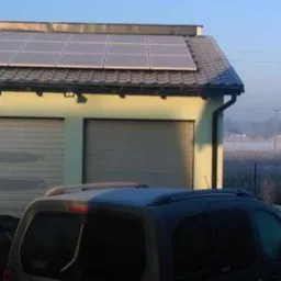 Panele fotowoltaiczne na dachu garażu dwustanowiskowego w zimowej scenerii, z oszronionymi samochodami na podjeździe i zamglonym polem w tle.