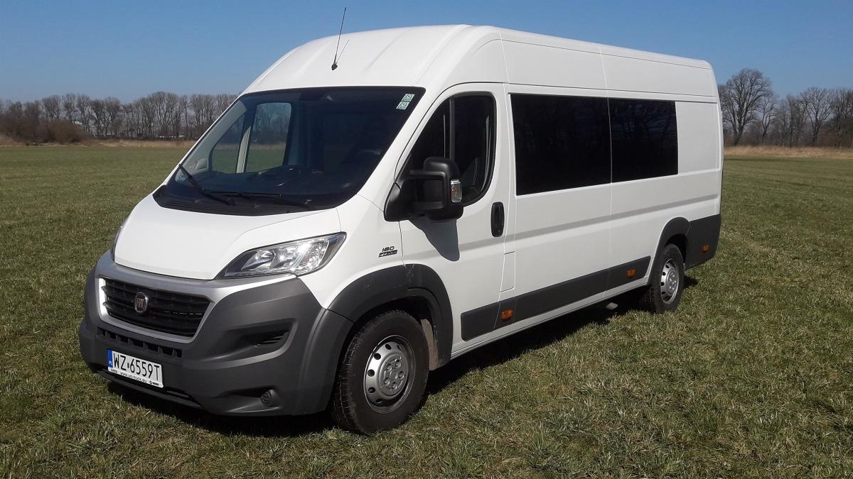 Biały Fiat Ducato 9-osobowy na trawiastym terenie z drzewami w tle, widok z przodu i boku, z widoczną tablicą rejestracyjną.