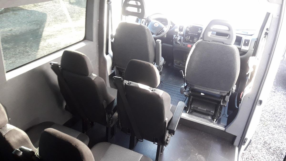 Wnętrze dziewięcioosobowego Fiata Ducato z widocznymi fotelami pasażerów, kierownicą i deską rozdzielczą, fotografowane z tyłu pojazdu.