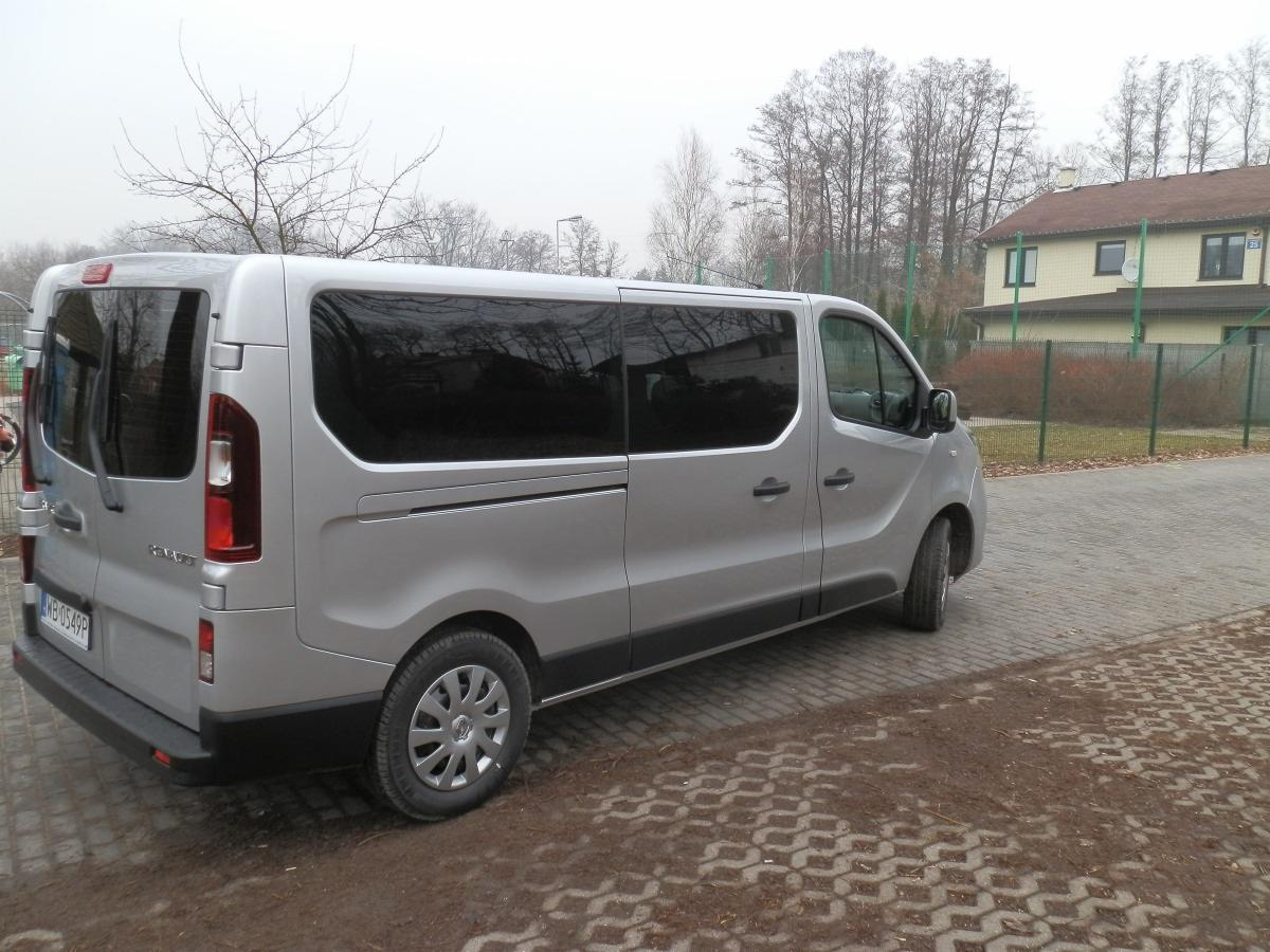 Srebrny Renault Trafic 9-osobowy zaparkowany na brukowanej nawierzchni, widok z tyłu i boku, w tle drzewa i budynek mieszkalny.