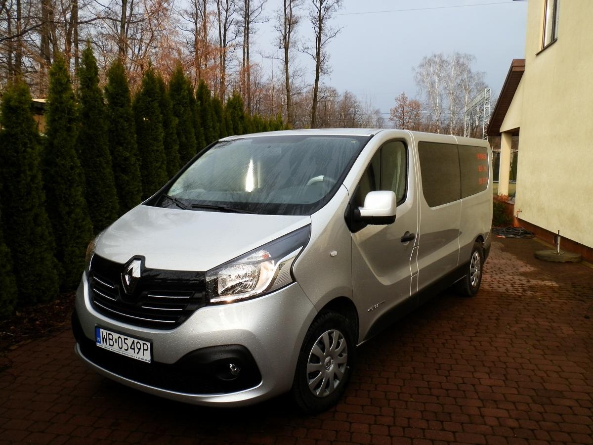 Srebrny Renault Trafic 9-osobowy na brukowanym podjeździe, z rzędem wysokich tuj w tle i fragmentem jasnego budynku po prawej stronie.