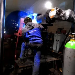 Spawanie elementu konstrukcji stalowej w warsztacie, widoczny spawacz w kombinezonie ochronnym, iskry i butla z gazem w tle.