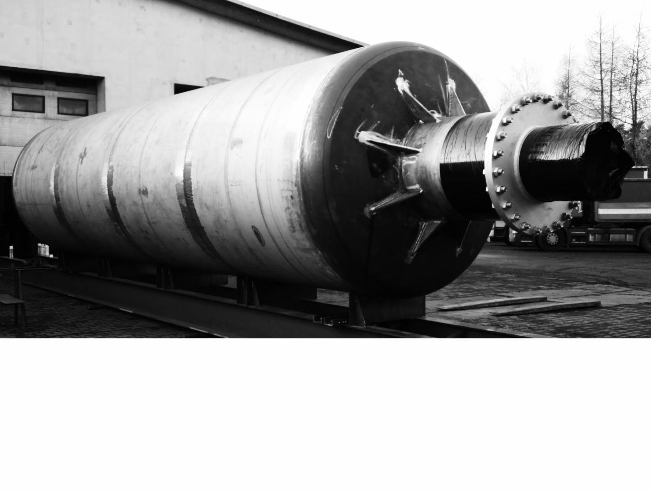 Czarno-białe zdjęcie dużego, poziomego zbiornika stalowego z widocznymi spawami, umieszczonego na metalowej konstrukcji, z fragmentem budynku w tle i ciężarówką w oddali.