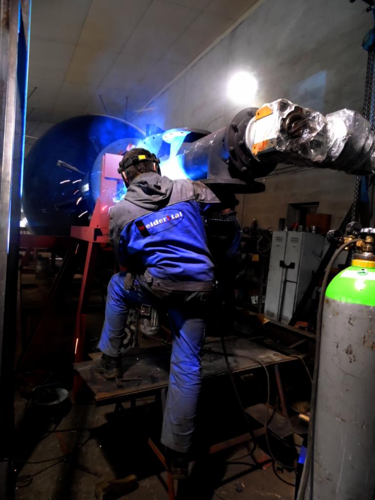 Spawanie elementu konstrukcji stalowej w warsztacie, widoczny spawacz w kombinezonie ochronnym, iskry i butla z gazem w tle.