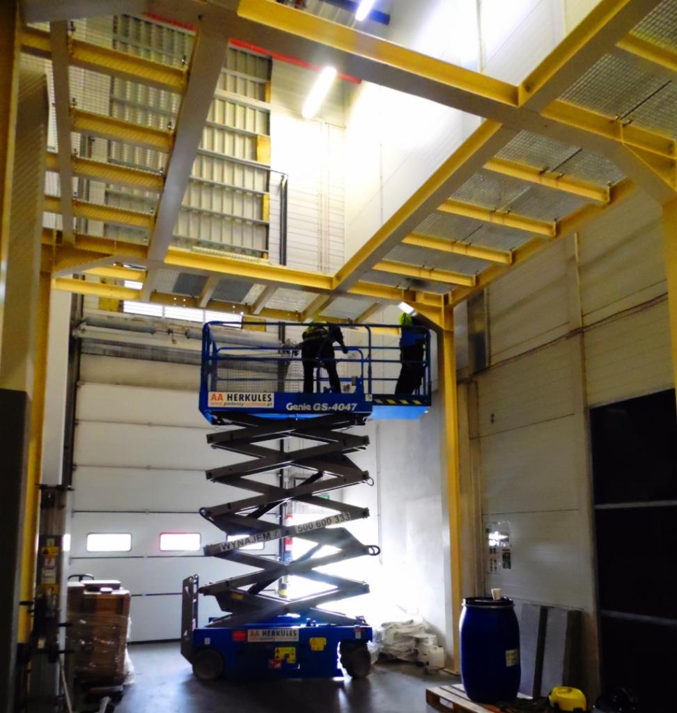 Prace montażowe na wysokości w hali magazynowej z wykorzystaniem podnośnika nożycowego, widoczne żółte elementy konstrukcji stalowej pod sufitem.