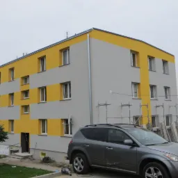 Trzypiętrowy budynek mieszkalny po odnowieniu elewacji, pomalowany na szaro i żółto, z widocznymi rusztowaniami i zaparkowanym samochodem osobowym.