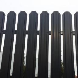 Dachy Kraśnik - Fragment nowoczesnego, metalowego ogrodzenia panelowego w kolorze grafitowym, z widocznymi elementami montażowymi, na tle budynku i drzew.