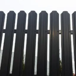 Fragment nowoczesnego, metalowego ogrodzenia panelowego w kolorze grafitowym, z widocznymi elementami montażowymi, na tle budynku i drzew.