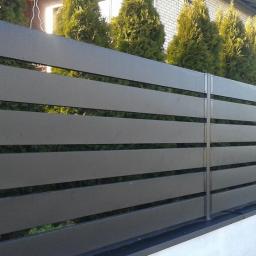 Dachy Kraśnik - Nowoczesne ogrodzenie panelowe z poziomych, szarych desek kompozytowych, widoczne na tle zielonych tuj i jasnego budynku.