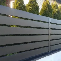 Nowoczesne ogrodzenie panelowe z poziomych, szarych desek kompozytowych, widoczne na tle zielonych tuj i jasnego budynku.