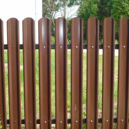 Dachy Kraśnik - Brązowe metalowe ogrodzenie palisadowe z widocznymi nitami, na tle zielonej trawy i krzewów.