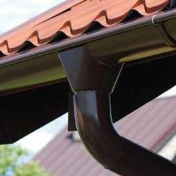 Dachy Kraśnik - Brązowy system rynnowy zamontowany na dachu z czerwonej dachówki, widok z dołu na tle błękitnego nieba.