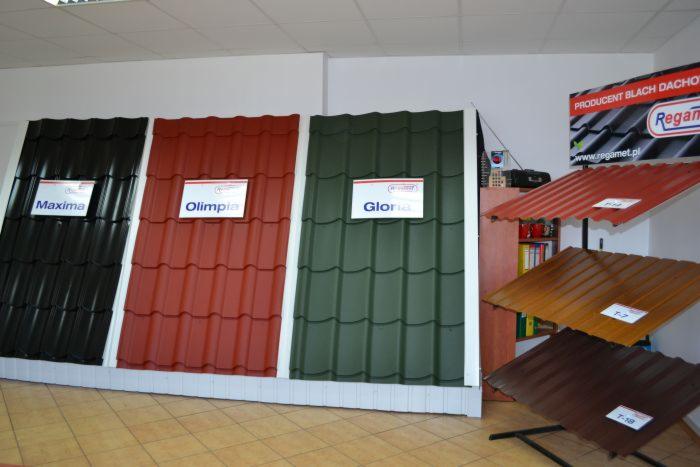 Ekspozycja próbek blachodachówki w różnych kolorach (czarny, czerwony, zielony) z nazwami modeli Maxima, Olimpia, Gloria, oraz mniejsze próbki na stojakach w odcieniach brązu i czerwieni...
