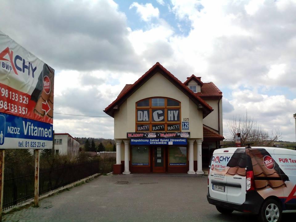 Budynek z napisem DACHY i reklamami, w tle błękitne niebo z chmurami, obok zaparkowany samochód z grafiką dachówki i logo firmy dekarskiej.