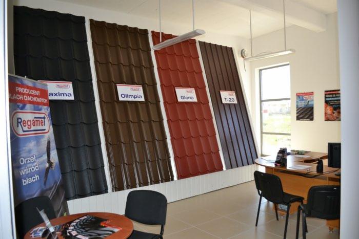 Ekspozycja paneli dachowych różnych kolorów i modeli (Axima, Olimpia, Gloria, T-20) w biurze firmy Regamel, z widocznym logo producenta na plakacie.