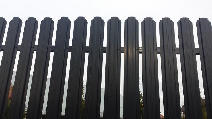 Fragment nowoczesnego, metalowego ogrodzenia panelowego w kolorze grafitowym, z widocznymi elementami montażowymi, na tle budynku i drzew.