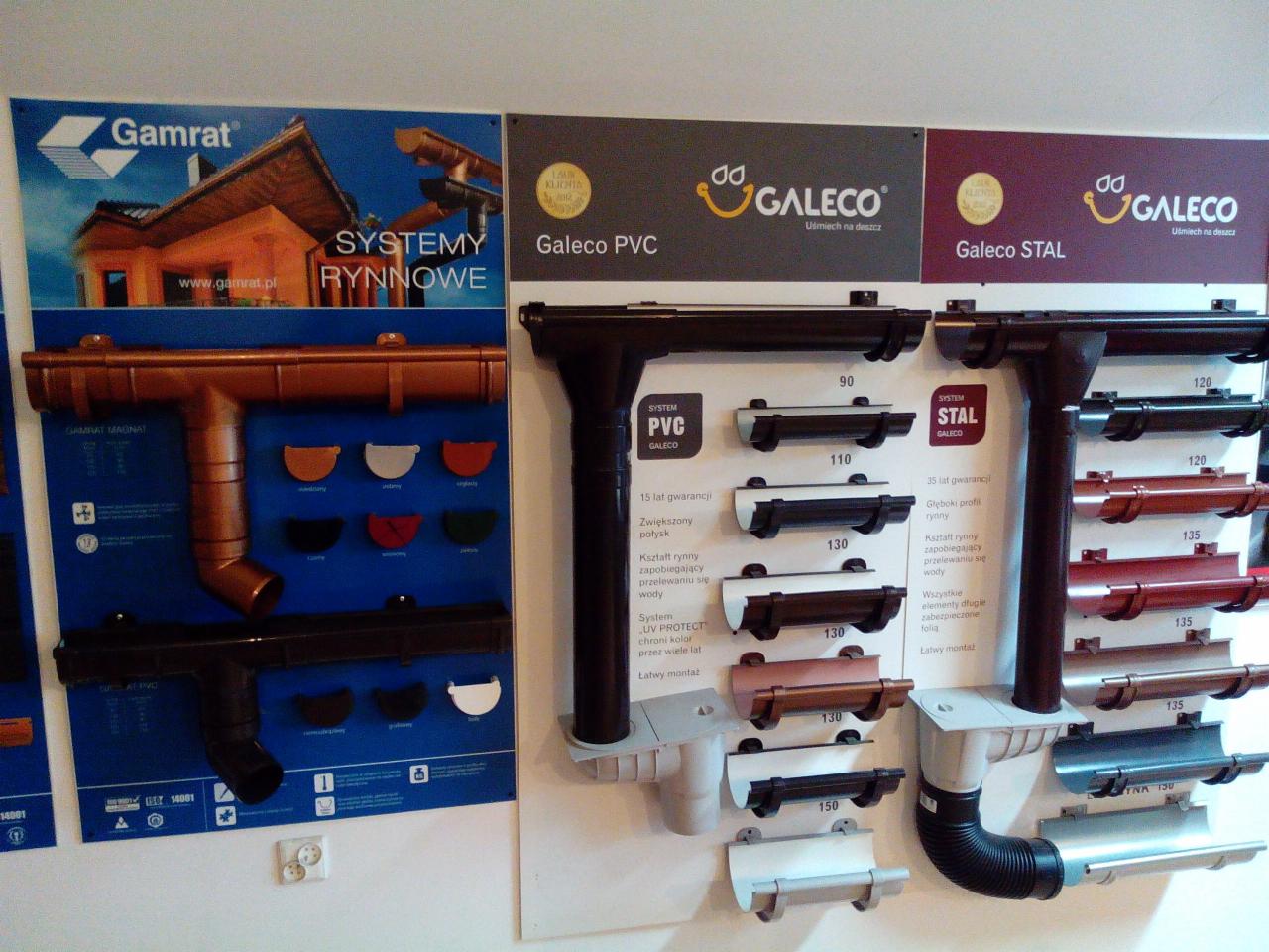 Ekspozycja systemów rynnowych różnych producentów, prezentacja przekrojów i kolorów, widoczne logotypy firm Gamrat i Galeco, materiały PVC i stal.