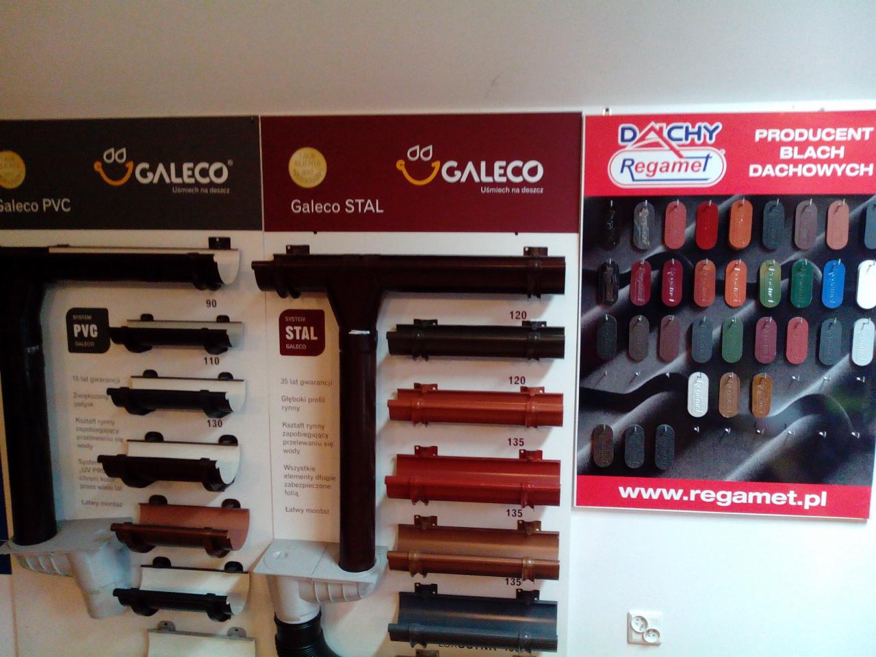 Ekspozycja systemów rynnowych PVC i stalowych marki Galeco oraz próbniki blach dachowych Regamet w punkcie sprzedaży w Kraśniku. Widoczne przekroje różnych modeli rynien w kolorach czarnym, białym...