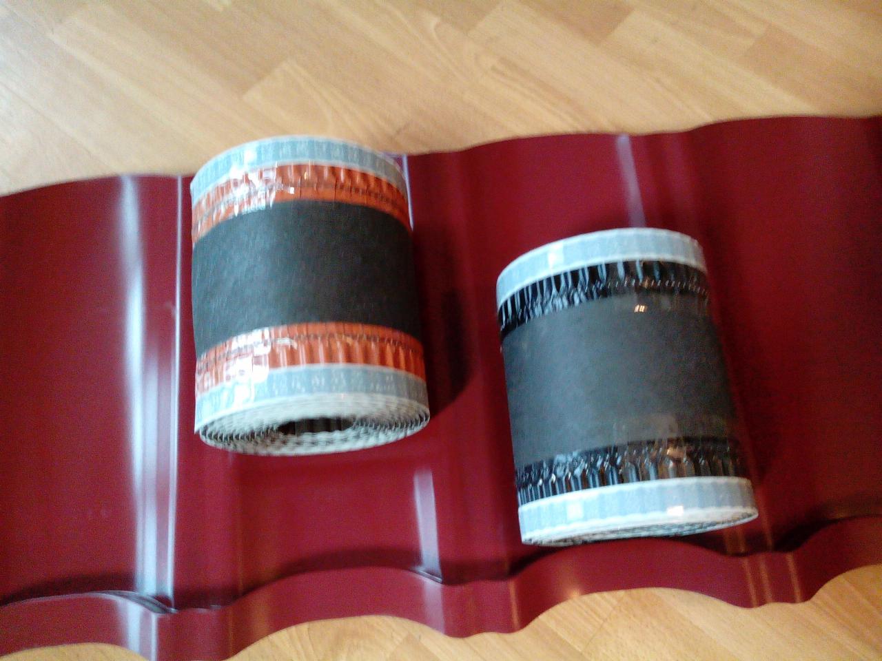 Dwa zwinięte pasy uszczelniające na arkuszu blachy dachowej w odcieniach czerwieni, leżące na drewnianej podłodze.