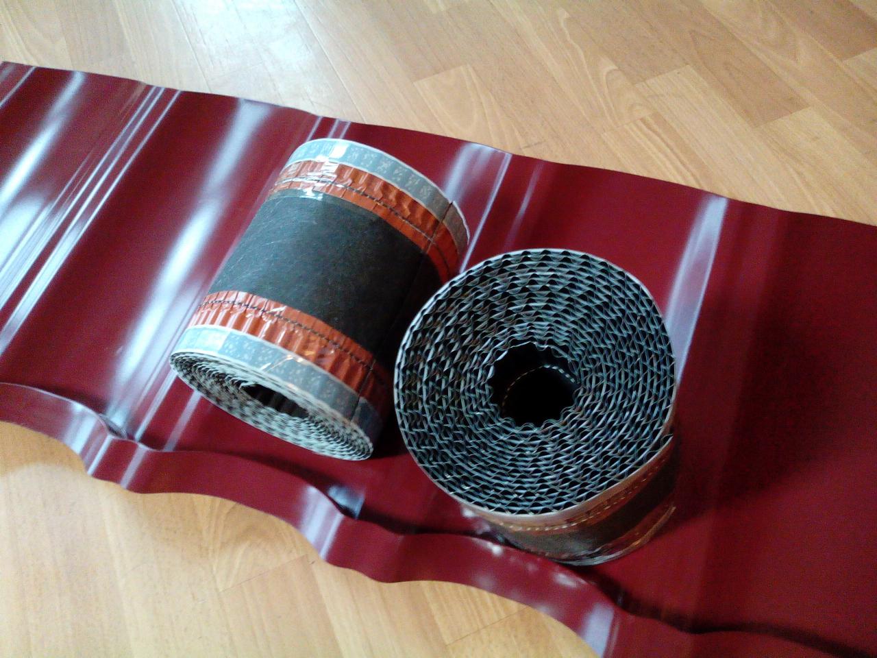Dwa zwinięte pasy uszczelniające do dachu, jeden z czarnym rdzeniem i srebrno-miedzianymi brzegami, leżą na arkuszu blachy dachowej w kolorze bordowym na drewnianej podłodze.
