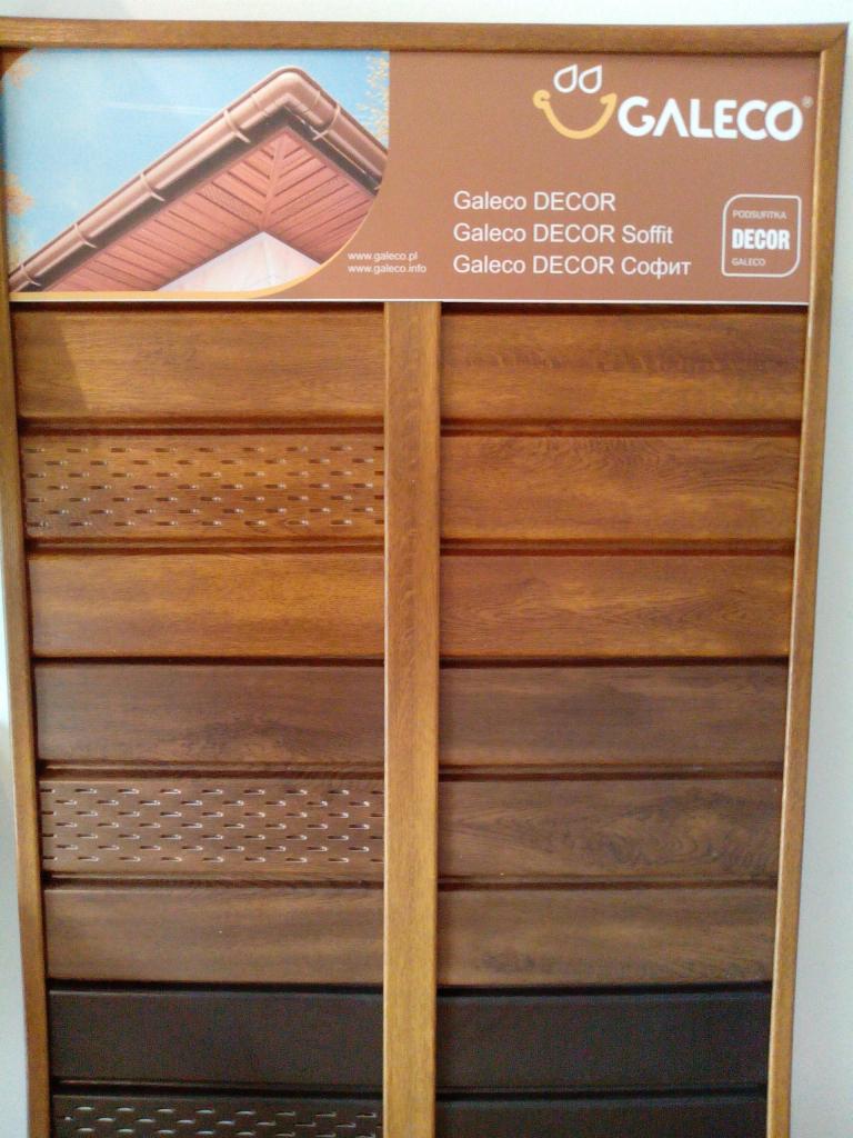 Ekspozycja próbek drewnianej podbitki dachowej Galeco Decor w różnych odcieniach brązu, z perforowanymi elementami wentylacyjnymi i górnym panelem z wizualizacją dachu z systemem rynnowym.