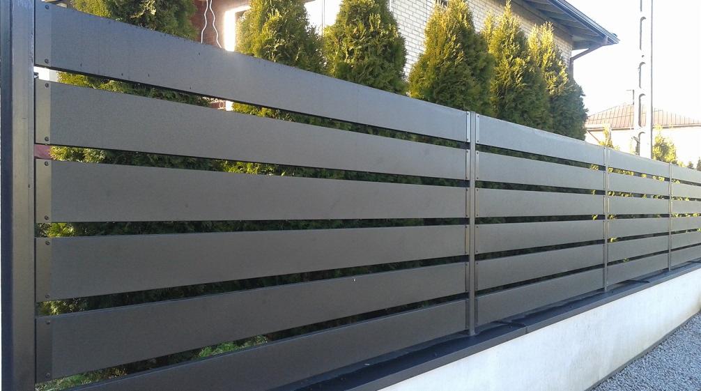 Nowoczesne ogrodzenie panelowe z poziomych, szarych desek kompozytowych, widoczne na tle zielonych tuj i jasnego budynku.