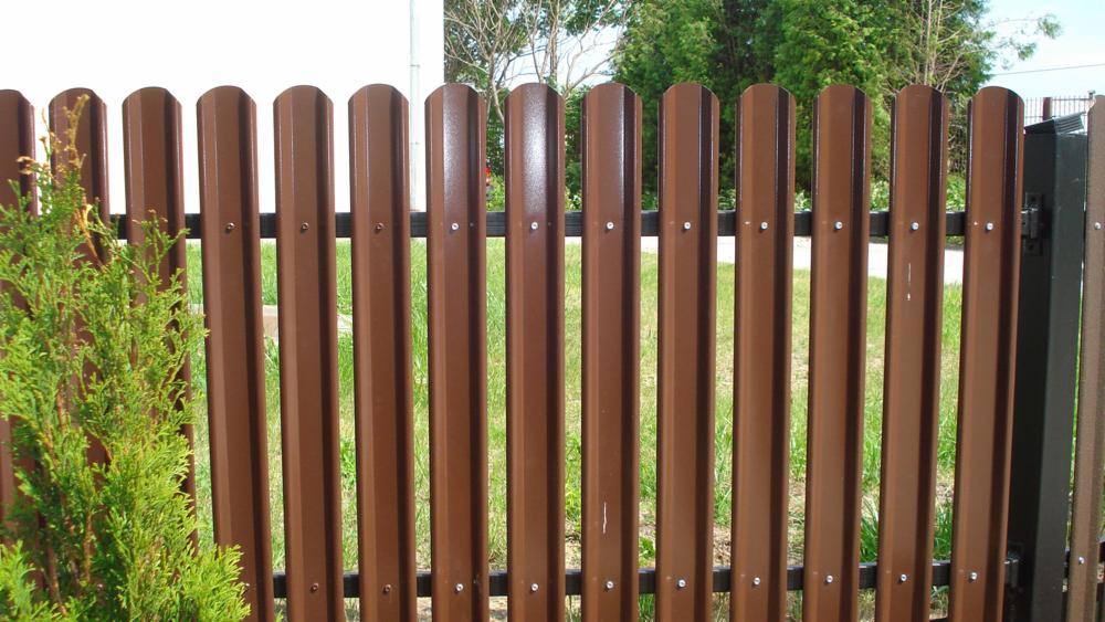 Brązowe metalowe ogrodzenie palisadowe z widocznymi nitami, na tle zielonej trawy i krzewów.