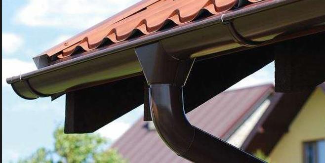 Brązowy system rynnowy zamontowany na dachu z czerwonej dachówki, widok z dołu na tle błękitnego nieba.