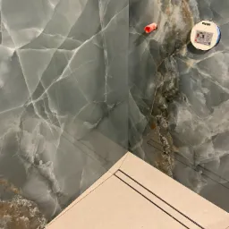 Fragment ściany wyłożonej płytami imitującymi marmur z widocznymi elementami instalacji wodnej i odpływem liniowym w podłodze.