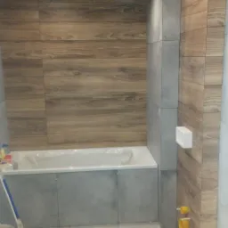 Łazienka w trakcie remontu, widoczne płytki imitujące drewno na ścianach, szare płytki na podłodze i obudowie wanny, narzędzie do cięcia płytek w lewym dolnym rogu, surowy stan instalacji...