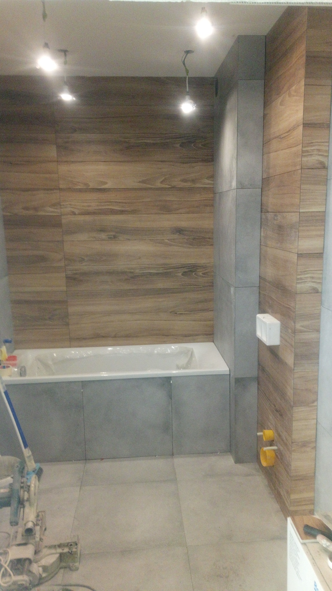 Łazienka w trakcie remontu, widoczne płytki imitujące drewno na ścianach, szare płytki na podłodze i obudowie wanny, narzędzie do cięcia płytek w lewym dolnym rogu, surowy stan instalacji...