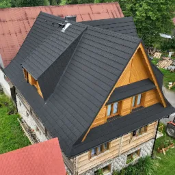 Nowy, ciemny dach z dachówki na drewnianym domu z kamienną podmurówką, widok z góry, w tle czerwony dach innego budynku i drzewa.