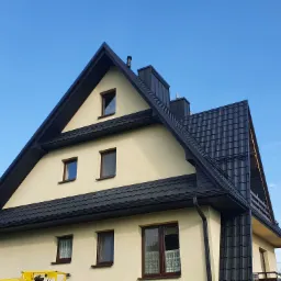 Dom jednorodzinny z ciemną, profilowaną blachą na dachu i kremową elewacją, widziany od dołu pod kątem, z fragmentem żółtego podnośnika widocznym w dolnym rogu. Widoczne okna z brązowymi ramami...