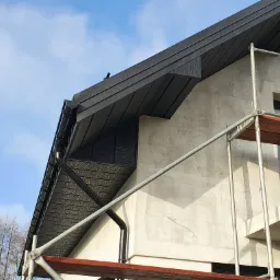 Montaż ciemnej podbitki dachowej na surowym budynku, widoczne rusztowanie i drabina, błękitne niebo w tle.