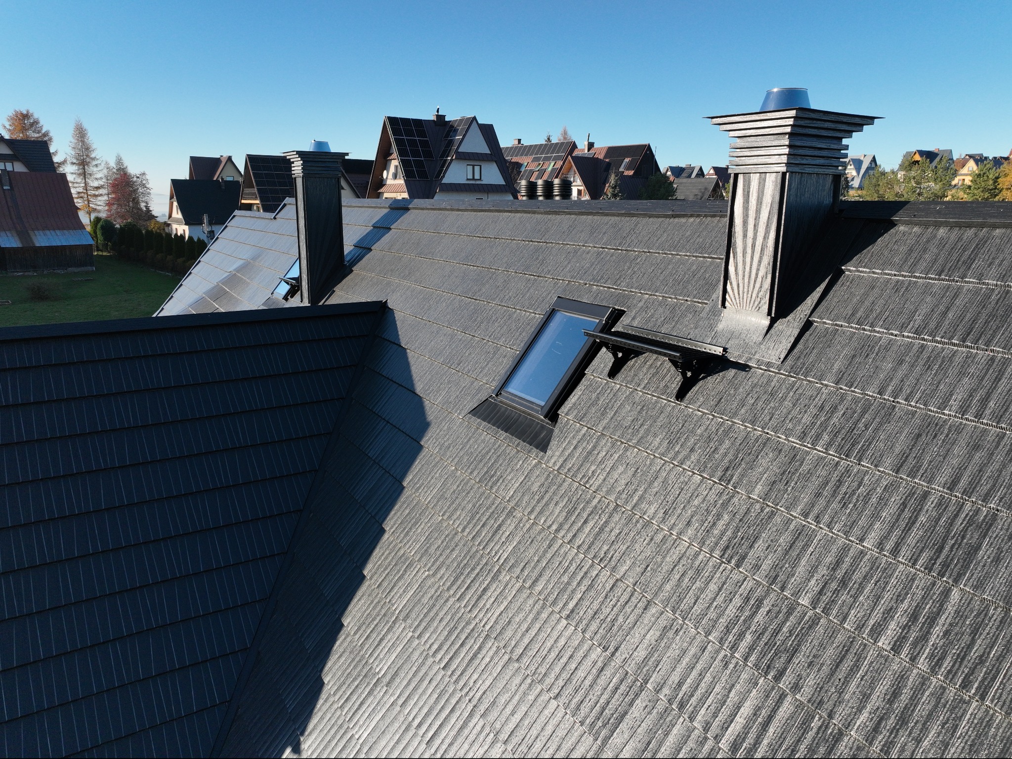 Szary dach z oknem dachowym i kominem na tle zabudowy jednorodzinnej w słoneczny dzień.