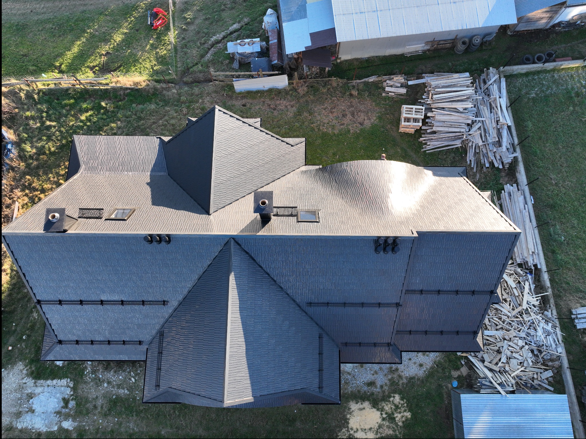 Widok z góry na ciemny dach z dachówki bitumicznej z kominami i oknami dachowymi, obok sterta tarcicy budowlanej, na trawiastym terenie.