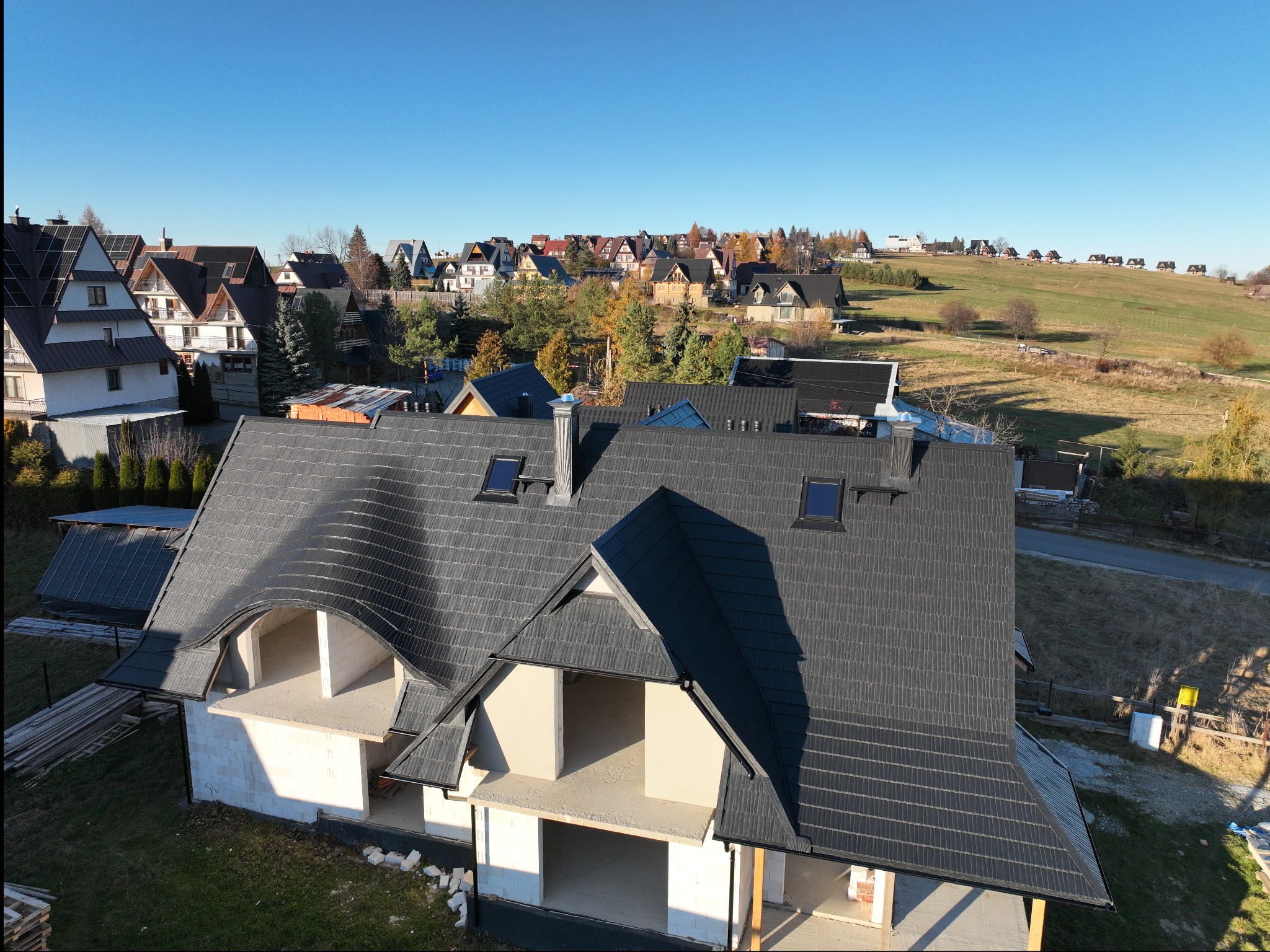 Dom w trakcie budowy z szarą dachówką karpiówką, widok z góry na osiedle domów i wzgórza w oddali, słoneczny dzień.