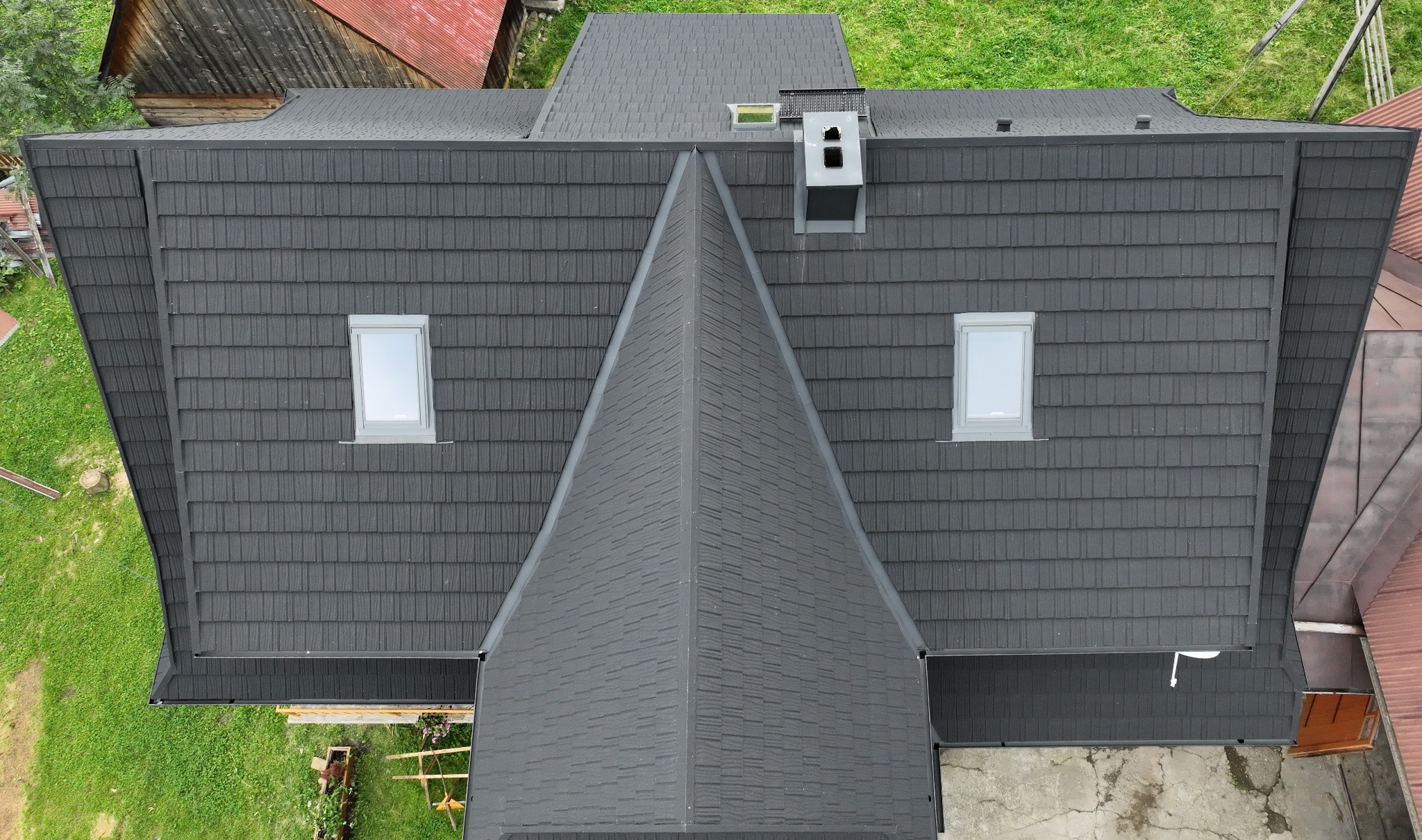 Widok z góry na ciemnoszary dach z dachówkami bitumicznymi, dwa okna dachowe i komin na tle zieleni i innych budynków.