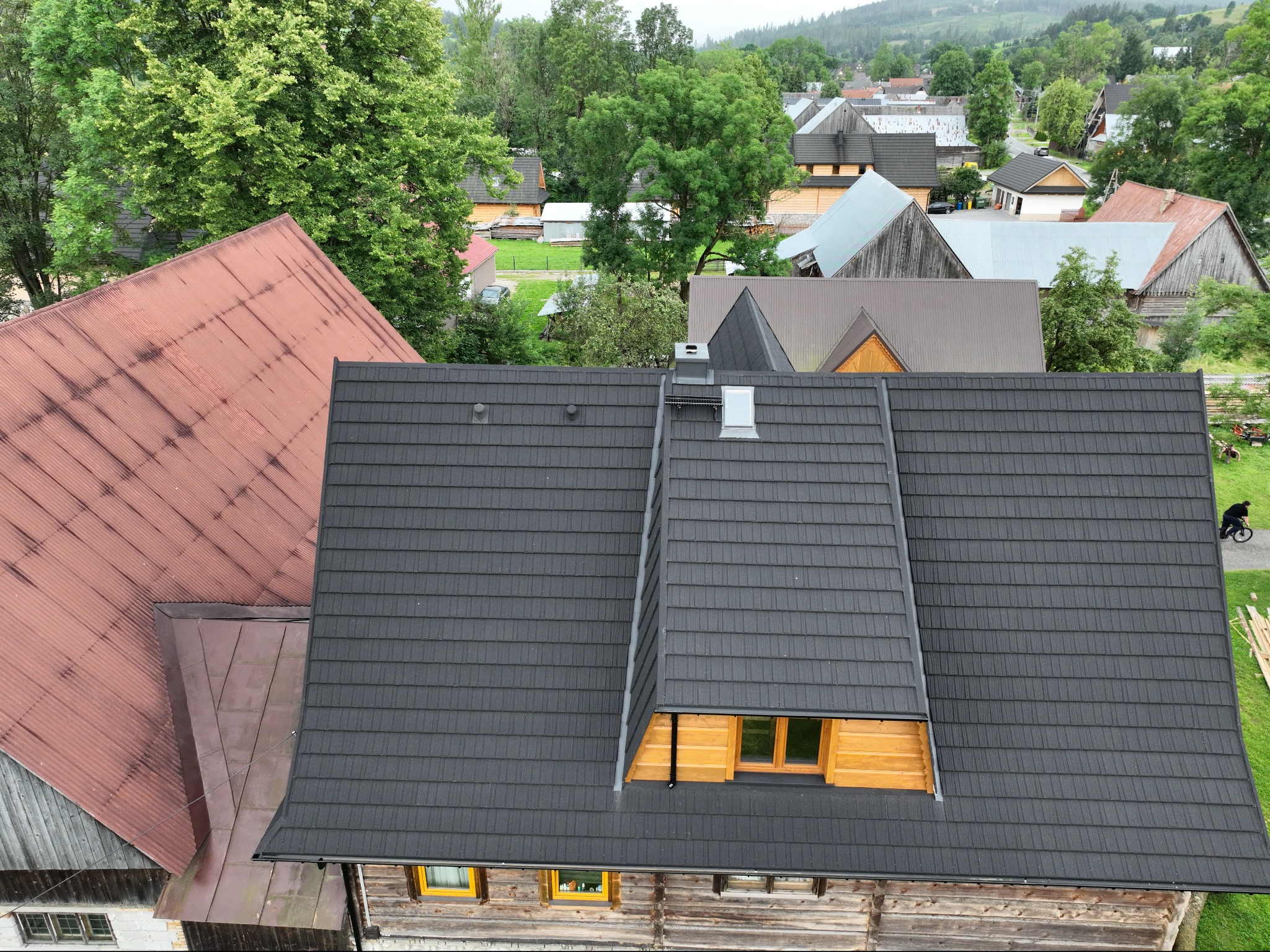 Widok z góry na dachy domów w górskiej miejscowości, z dominującym ciemnoszarym dachem z nową lukarną, obok dachy z blachy falistej w odcieniach czerwieni i brązu, w tle zalesione wzgórza...