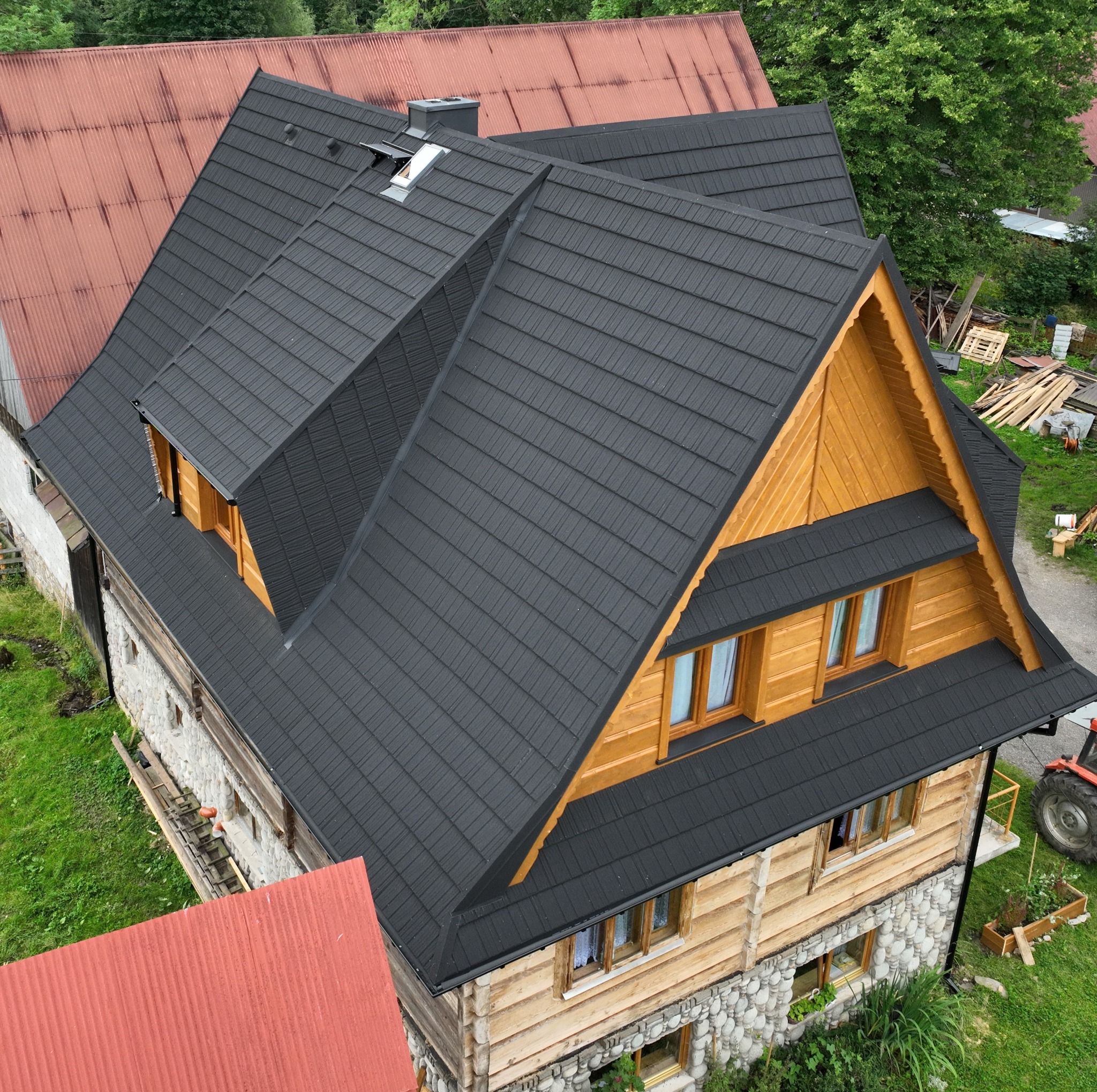 Nowy, ciemny dach z dachówki na drewnianym domu z kamienną podmurówką, widok z góry, w tle czerwony dach innego budynku i drzewa.