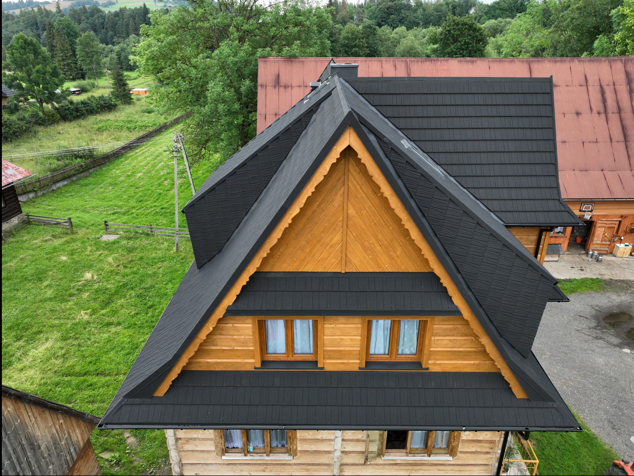 Widok z góry na dom z bali z ciemnoszarą dachówką modułową, widoczne drewniane elementy wykończeniowe i okna z firankami, w tle zieleń.