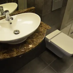 Nowoczesna łazienka z owalną umywalką nablatową na blacie z brązowego marmuru, podwieszana toaleta i szara glazura w kabinie prysznicowej.