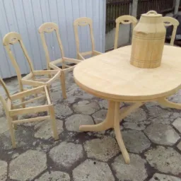 Stół owalny i cztery krzesła z jasnego drewna, po obróbce, ale przed malowaniem, stojące na kamiennej posadzce; na stole stoi drewniana beczułka.