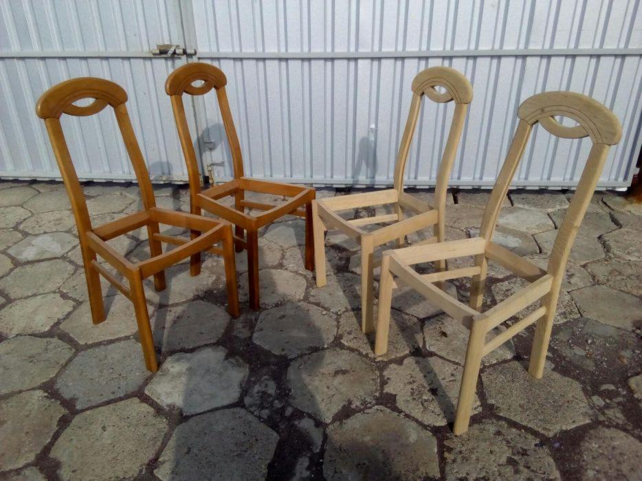 Cztery niedokończone drewniane krzesła z charakterystycznym wycięciem w oparciu, dwa w kolorze naturalnego drewna, a dwa w odcieniu brązu, stojące na kamiennej posadzce w Krotoszynie. Przygotowanie...